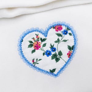 Può includere: Una toppa a forma di cuore con un motivo floreale rosa, blu e giallo su sfondo bianco. La toppa è bordata da pizzo blu ed è su un tessuto bianco.