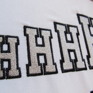 May include: Embroidered letters spelling out "HHHH" in a white, grey, and black colour scheme. The letters are stitched in a bold, blocky font.