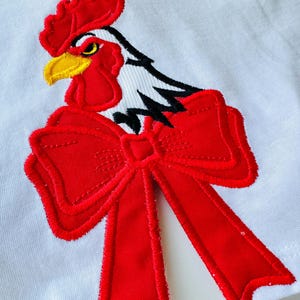 Peut inclure: T-shirt blanc avec un motif brodé d'un coq rouge, bec jaune et grand nœud rouge. Le coq a des détails noirs et blancs. Le motif est centré sur le t-shirt, offrant un look audacieux.