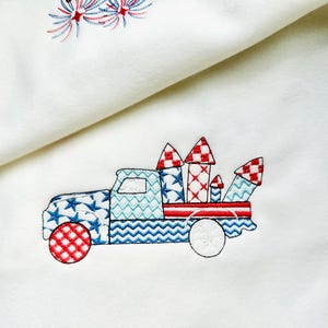 Op de afbeelding: Geborduurd ontwerp van een patriottische vrachtwagen met vuurwerk. De vrachtwagen heeft rood, wit en blauwe patronen, waaronder sterren en ruitjes. De wielen zijn wit en de achtergrond is een zachte witte stof.