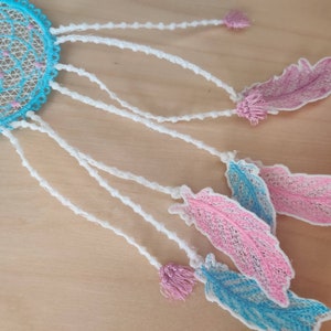May include: A blue and white dreamcatcher with pink feathers hanging from it. The dreamcatcher is made of lace and has a beaded border. The feathers are also made of lace and have a delicate, feminine look.
