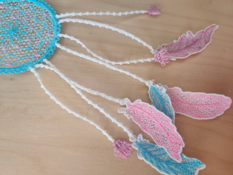May include: A blue and white dreamcatcher with pink feathers hanging from it. The dreamcatcher is made of lace and has a beaded border. The feathers are also made of lace and have a delicate, feminine look.