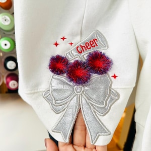 May include: White sweatshirt with a silver bow and pom-poms embroidered on the cuff. The pom-poms are red and purple. The bow has the word "cheer" embroidered on it in red.