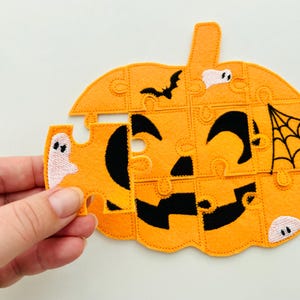 May include: Orange felt Halloween puzzle in the shape of a jack-o'-lantern. The puzzle features a black face, a spider web, and a bat. A hand holds a puzzle piece with a ghost.