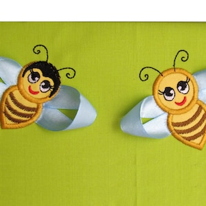 May include: Two embroidered bee appliques with black and brown stripes, yellow faces, and big eyes. The bees are attached to light blue ribbon bows.
