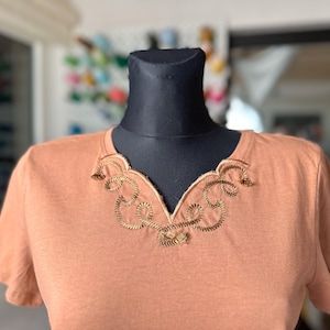 Könnte beinhalten: Ein pfirsichfarbenes T-Shirt mit einem goldenen, gestickten Ausschnitt mit einem wirbelnden Design.