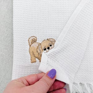 May include: White waffle-weave towel with a tan and brown embroidered puppy design. The towel has a fringe along the bottom edge. The puppy is facing away from the viewer, with its head turned to the side.