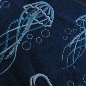 May include: Embroidered blue fabric with a design of three white jellyfish and white bubbles. The jellyfish have a bell-shaped head and long, flowing tentacles.