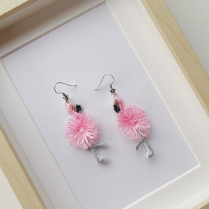 May include: A pair of pink flamingo earrings with silver wire and black accents. The earrings are displayed in a white frame.
