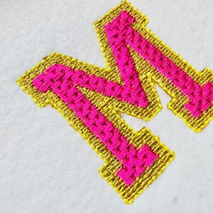 May include: A pink letter M with a gold outline. The letter is embroidered on a white fabric.