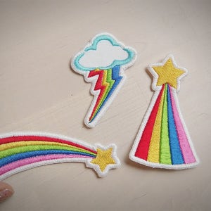May include: Three embroidered iron-on patches featuring rainbow designs. One patch is a rainbow shooting star, another is a rainbow cloud with a lightning bolt, and the third is a rainbow shooting star.