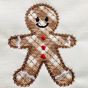 May include: Embroidered gingerbread man design on a white fabric background. The gingerbread man is brown with white and red accents. The gingerbread man has a smiling face.