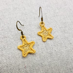 May include: A pair of yellow star-shaped earrings with bronze-colored hooks. The earrings are made of a delicate, open-weave material, and are displayed on a light gray textured surface. The earrings are a jewelry item.