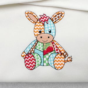 May include: An embroidered patch featuring a stylized donkey character composed of various colorful fabric patterns. The design includes orange chevron, blue gingham, green polka dots, and red zigzag motifs on a white fabric background.