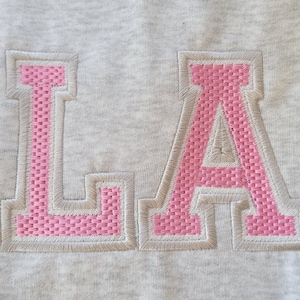 May include: A gray sweatshirt with the letters "LA" embroidered in pink.