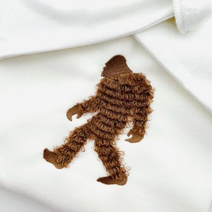 May include: White fabric with a brown embroidered Bigfoot design. The Bigfoot is depicted walking with detailed fur-like texture. The design is centered on the fabric, with the white background providing contrast.