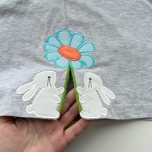 Può includere: Una maglietta grigia con un fiore blu e due coniglietti bianchi ricamati. Il fiore ha un centro arancione e uno stelo verde. I coniglietti si guardano e hanno orecchie lunghe.