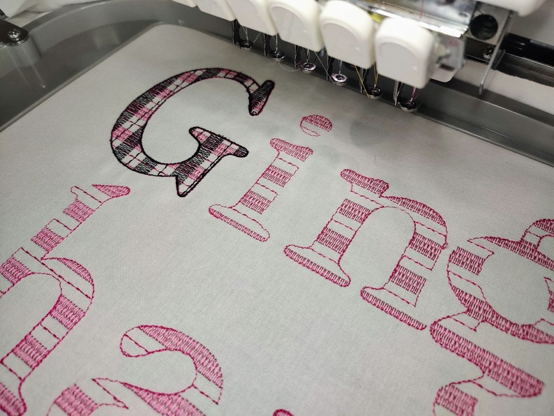Pode incluir: Um close-up de uma m&aacute;quina de bordado costurando o nome "Ginger" em fio rosa e preto em um tecido branco. As letras est&atilde;o em uma fonte estilizada com um padr&atilde;o xadrez.