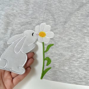 Può includere: Un toppa di tessuto grigio con un coniglio bianco ricamato che annusa una margherita bianca con un centro giallo e uno stelo verde.