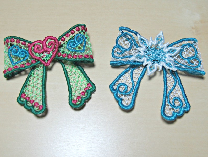 May include: Two embroidered lace bows, one green with pink hearts and one blue with a white snowflake.