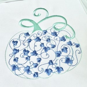 May include: Embroidered design of a pumpkin with blue and green leaves and vines. The design is on a white fabric.