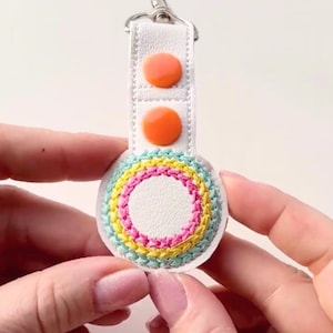 May include: A white leather keychain with a round, embroidered circle. The circle has a white center and three concentric rings of turquoise, yellow, and pink embroidery. The keychain has two orange buttons.