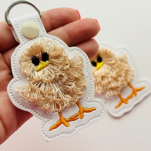 May include: Two white felt keychains with embroidered fluffy yellow chicks. The chicks have black eyes and orange beaks.