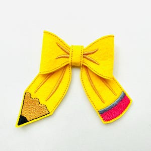 May include: A yellow felt bow resembling a pencil, featuring brown and black embroidery on the pencil tip and a red, grey, and blue band. This school-themed accessory is designed as a bow.