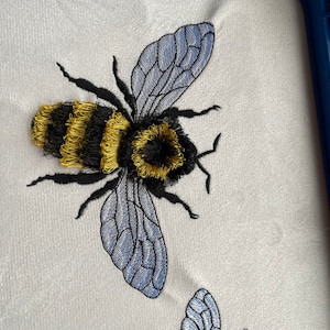 May include: Embroidered design of two bumblebees with black and yellow stripes and blue wings on a white fabric background.