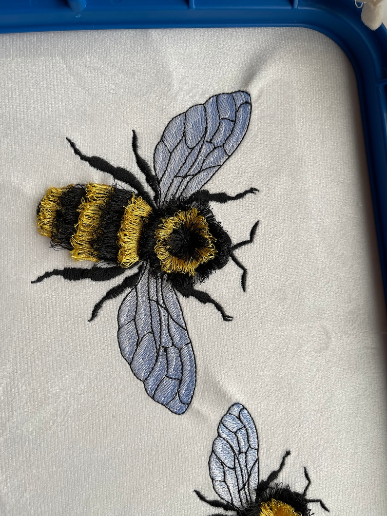 May include: Embroidered design of two bumblebees with black and yellow stripes and blue wings on a white fabric background.