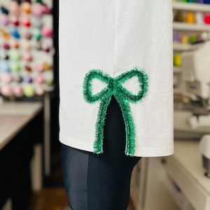 May include: White t-shirt with a green fuzzy bow sewn on the side seam.
