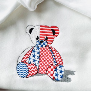May include: Embroidered teddy bear design in red, white, and blue on a white background. The bear features a patriotic design with stars and stripes, reminiscent of the American flag. The bear is sitting with its legs crossed.