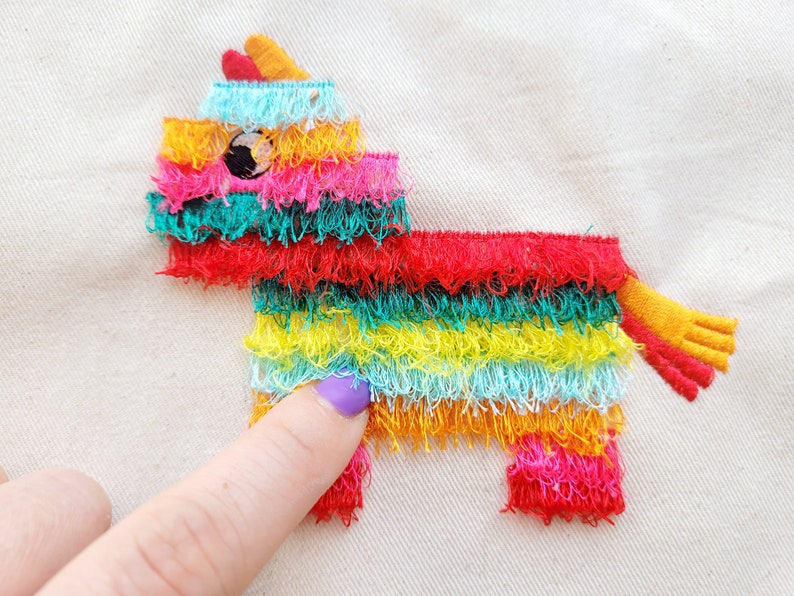 Puede incluir: Una pi&ntilde;ata bordada de colores con forma de burro con un borde de flecos. La pi&ntilde;ata est&aacute; hecha de tela y tiene un esquema de colores rojo, amarillo, verde, azul y rosa.