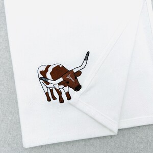 May include: White folded towel with an embroidered image of a brown and white longhorn steer. The steer is facing left with its horns curved upwards. The towel is made of a soft, white fabric.