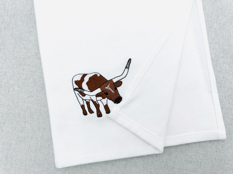 May include: White folded towel with an embroidered image of a brown and white longhorn steer. The steer is facing left with its horns curved upwards. The towel is made of a soft, white fabric.