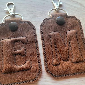 Puede incluir: Dos llaveros de cuero marrón con anillas metálicas y cierres de presión. Los llaveros presentan las letras "E" y "M" cosidas en relieve.
