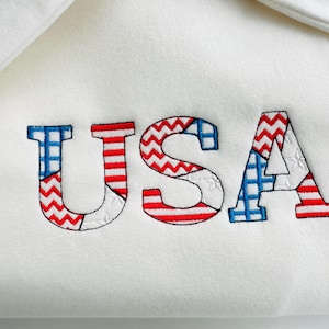 Könnte beinhalten: Weißer Stoff mit dem Wort "USA" in roter, weißer und blauer Patchwork-Art gestickt.