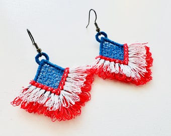 May include: A pair of handmade earrings featuring a square blue and red woven design with white and red fringe. The earrings have a hook closure and are set against a white background.