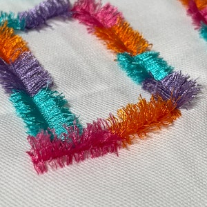 Op de afbeelding: Een close-up van een witte stof met twee kleurrijke vierkanten van gefranst stof. De vierkanten zijn gemaakt van oranje, roze, paars en turquoise stof.