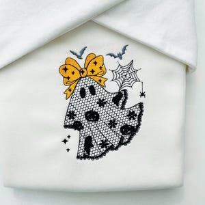May include: White fabric with an embroidered Halloween ghost design. The ghost is made of black lace and has a yellow bow. A spider web, bats, and stars complete the spooky design.