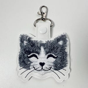 May include: White key chain with a gray and white embroidered cat face. The cat has a white nose and whiskers and is smiling.