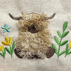 May include: Embroidered design of a Highland cow with long, shaggy fur, brown horns, and hooves. The cow is surrounded by embroidered yellow and blue flowers and green leaves on a beige fabric background.