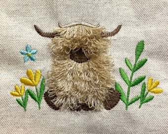 May include: Embroidered design of a Highland cow with long, shaggy fur, brown horns, and hooves. The cow is surrounded by embroidered yellow and blue flowers and green leaves on a beige fabric background.