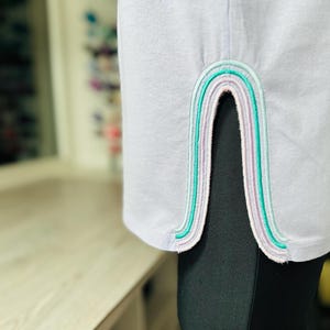 May include: A lavender colored shirt with a rainbow-colored trim on the side seam.