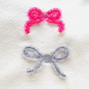 Op de afbeelding: Twee geborduurde strikken op een witte stoffen achtergrond. De bovenste strik is roze en de onderste strik is grijs.