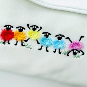 Puede incluir: Tela blanca con un diseño bordado de seis ovejas coloridas. Las ovejas tienen cuerpos esponjosos en rojo, amarillo, azul y rosa. Cabezas y patas negras, con detalles de hierba verde.
