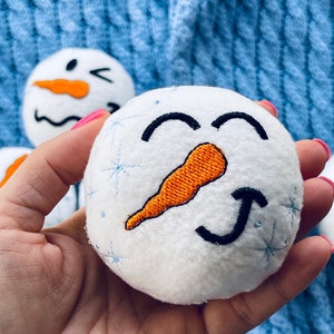 May include: A white plush snowman ornament with a smiling face, orange carrot nose, and black eyes. The snowman is embroidered with snowflakes and is being held in a hand.