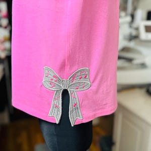 May include: A pink t-shirt with a gray bow embroidered on the sleeve. The bow is decorated with pink musical notes.