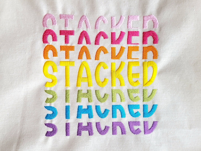 May include: A white fabric with the word "STACKED" embroidered in rainbow colors. The word is repeated five times, with each repetition in a different color: pink, red, orange, yellow, green, blue, and purple.