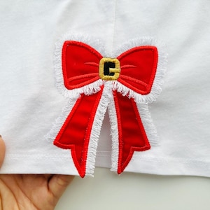 May include: A white fabric with a red embroidered bow with a gold buckle. The bow has a white fringe around the edges.